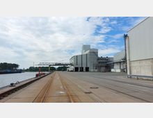 Industriegebiet an einem Hafen mit einem großen, grauen Lagergebäude und Schienen auf dem Betonboden. Rechts befinden sich weitere Lagerstrukturen, links ist das Wasser mit einem Anleger und bewaldete Bereiche im Hintergrund sichtbar.