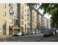 Straßenszene mit modernen Wohn- und Geschäftshäusern aus gelbem Backstein, gekennzeichnet durch große Fenster und runde Fensterelemente. Am Straßenrand stehen parkende Autos, während im Erdgeschoss mehrere Geschäfte und Restaurants zu sehen sind.