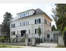 Zweistöckiges weißes Wohnhaus mit schwarzem Schieferdach und Fensterläden. Vor dem Haus befinden sich ein Zaun mit Tor, ein gepflasterter Weg und umgebende Bepflanzung, einschließlich eines großen Baumes rechts.