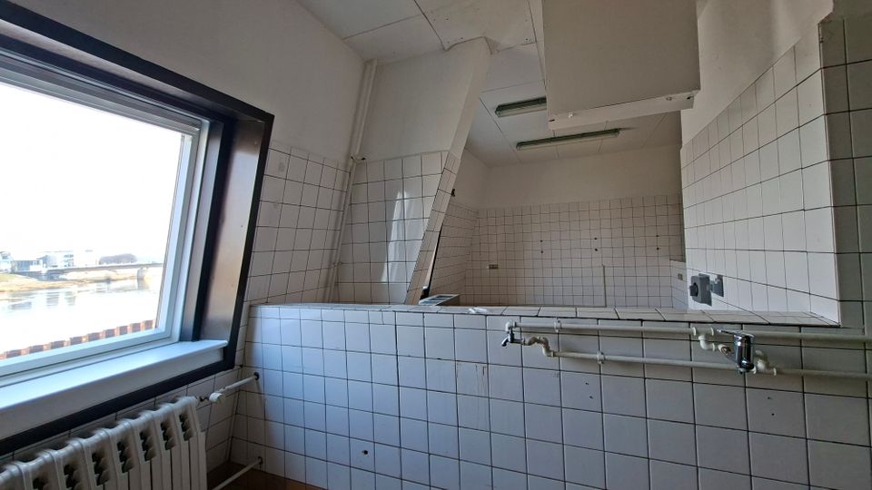 Innenraum einer ehemaligen Großküche oder eines Arbeitsraums mit weiß gefliesten Wänden, großen Abluftkästen unter der Decke und sichtbaren Rohrleitungen über einer gefliesten Trennwand. Links befindet sich ein großes Fenster mit Blick nach draußen und darunter ein Heizkörper.