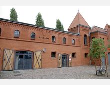 Backsteinbau mit einem historischen Industriecharakter, gekennzeichnet durch Rundbogenfenster, hölzerne Doppeltüren und ein Satteldach mit kleinen Türmchen. Im Vordergrund Kopfsteinpflaster und ein Baum sowie ein geparktes Fahrrad.