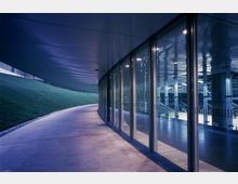 Gebogenes Außengelände mit einer asphaltierten, überdachten Gehwegfläche, flankiert von einer Glasfassade auf der rechten Seite, die den Blick ins Innere eines beleuchteten modernen Gebäudes freigibt, sowie einer begrünten Böschung auf der linken Seite.