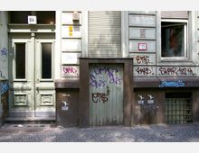 Fassade eines älteren Gebäudes mit einer grünen Eingangstür und einer kleineren Holztür daneben, beide mit Graffiti besprüht. Das Gebäude hat helle Wandvertäfelungen, heruntergelassene Rollläden und ein teils geöffnetes Fenster; der Gehweg besteht aus Kopfsteinpflaster.