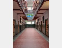 Innenansicht eines ehemaligen Gefängnisses mit einer roten Bodenfläche, Holztüren an beiden Seiten der Gänge und Metallgittern an der Decke. Im Hintergrund sind Fenster und ein Heizkörper zu sehen, die Tageslicht in den Raum lassen.
