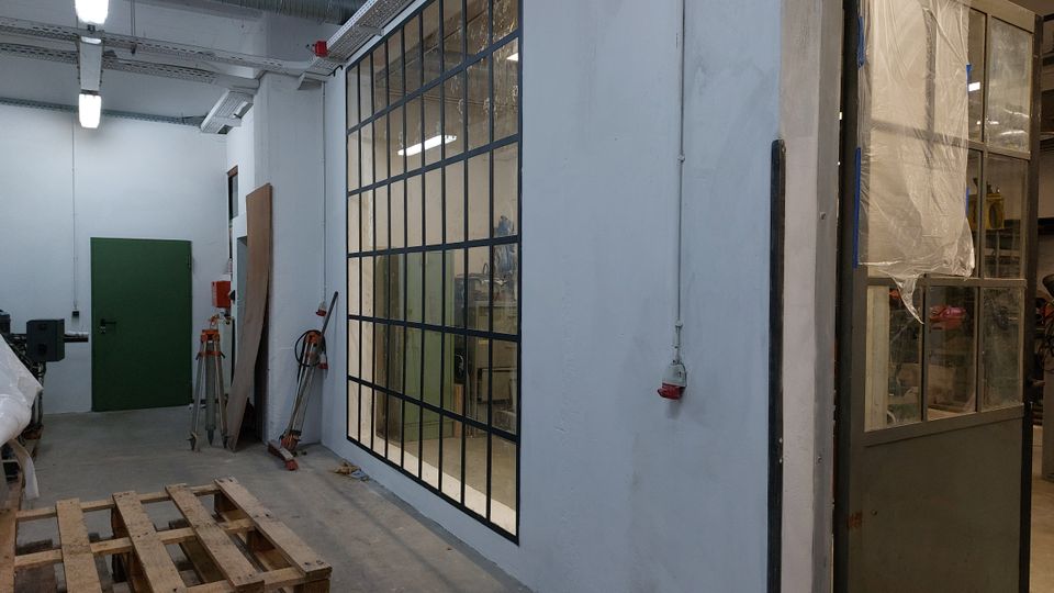 Industriehalle mit heller Beleuchtung, einer grünen Tür an der weißen Wand und einer großen Glaswand mit schwarzen Metallrahmen, die in einen angrenzenden Arbeitsbereich blicken lässt. Im Vordergrund ist eine Holzpalette, an den Wänden hängen Werkzeuge und Elektroinstallationen.