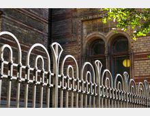 Detailansicht einer historischen Backsteinfassade mit verzierten Fensterbögen und kunstvollen Eisenzaunelementen im Vordergrund. Im oberen Bereich der Fassade sind dekorative Steinornamente sichtbar, während im Hintergrund ein gelbes Licht durch eines der Fenster scheint.