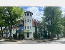 Eckgebäude mit weißer Fassade und einem hohen, turmähnlichen Erker, kombiniert mit roten Dachziegeln und kleinen Balkonen. Im Vordergrund sind Bäume, ein Gehweg, ein Parkplatzschild sowie eine Straßenecke mit Kopfsteinpflaster zu sehen.