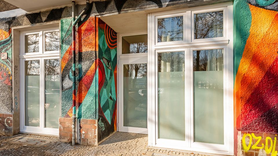 Eingangsbereich eines Gebäudes mit großen weißen Fenster- und Türelementen sowie teils blickdicht folierten Scheiben. Die Fassade ist mit farbenfrohem Graffiti und geometrischen Mustern gestaltet; davor liegt ein gepflasterter Gehweg mit Fallrohren an der Wand.