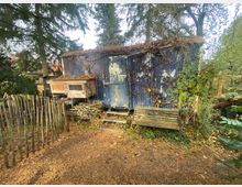 Ein blauer, verwitterter Bauwagen mit abblätternder Farbe steht in einem bewaldeten Garten. Daneben befinden sich eine Sitzbank, ein kleines Holzhäuschen mit Drahtgeflecht und ein Zaun aus Holzpfählen, umgeben von Bäumen und dichter Vegetation.
