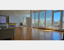 Heller Seminarraum mit großen Fensterfronten und Lamellenjalousien, die viel Tageslicht einlassen, ausgestattet mit Holzfußboden, einer Leinwand, einem Whiteboard und einigen Stühlen. Im Hintergrund ist ein Zugang zu einem Balkon mit Pflanzenkübeln zu sehen.