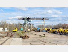 Ein Industriehof mit Schienennetz, großen Kränen und gelben Baustellenfahrzeugen. Auf der rechten Seite sind gelagerte Maschinenteile und Materialien, im Hintergrund stehen Bäume unter einem teils bewölkten Himmel.