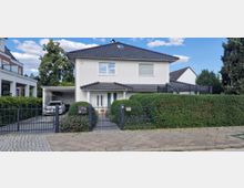 Zweigeschossiges Einfamilienhaus mit weißer Fassade und dunklem Walmdach, umgeben von einer hohen Hecke. Im Vorgarten befindet sich ein gepflasterter Weg und ein Carport mit einem geparkten Auto, alles eingefasst durch einen schwarzen Metallzaun.