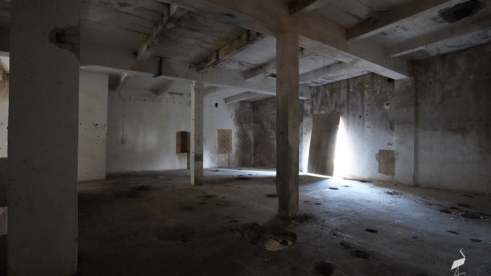 Großer, leerer Betonraum in einem verlassenen Industriegebäude mit massiven Stützpfeilern, niedriger Decke und fleckigen, abgenutzten Wänden. Seitlich fällt helles Tageslicht durch eine schmale Öffnung ein und wirft Schatten auf den staubigen Boden mit mehreren runden Vertiefungen.