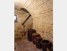 Ziegelgewölbter Kellerraum mit schmalen Treppenstufen, einer Mauer aus grobem Backstein und mehreren großen Keramiktöpfen entlang der Wand. Ein kleines Fenster mit Gitter bringt Licht in den rustikalen Raum.