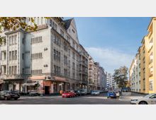 Straßenszene in einer Stadt mit mehrstöckigen Wohnhäusern im klassischen Stil auf der linken Seite und moderneren Gebäuden auf der rechten Seite; geparkte Autos säumen die Straße, und im Erdgeschoss befindet sich ein Restaurant mit dem Namen "Villa Italiana".