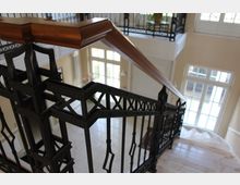 Treppenhaus mit geschwungener Treppe, die aus hellen Marmorstufen besteht, sowie einem dekorativen Geländer aus schwarzem Metall mit Holzhandlauf. Im Hintergrund sind hohe Fenster mit weißem Rahmen sichtbar, die Tageslicht in den Raum lassen.