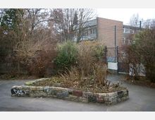 Ein urbaner Hof mit einem kleinen, rechteckigen Blumenbeet aus gemauerten Natursteinen, umgeben von kargen Büschen und Bäumen im Winter. Im Hintergrund sind eine Backsteinfassade eines Gebäudes sowie ein Zaun zu sehen.