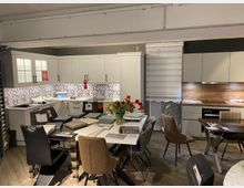 Ein Ausstellungsraum mit einer modernen Küchenzeile links und rechts, ausgestattet mit weißen und grauen Schränken, Holzelementen und einer Arbeitsplatte mit Dekorfliesen. Im Vordergrund stehen mehrere Esstische mit verschiedenen Stuhlmodellen, teilweise mit Mustern und Informationsmaterial belegt.