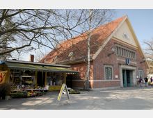 Backsteinbau im traditionellen Baustil mit Satteldach, bezeichnet als "Bhf. Oskar-Helene-Heim", mit einem Blumenladen im Vordergrund. Der Platz vor dem Gebäude ist gepflastert, und kahle Baumäste rahmen die Szene ein.