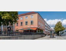 Eckgebäude mit orangefarbener Fassade, in dem sich im Erdgeschoss die „Mocca Milch Eisdiele“ mit markisenüberdachter Terrasse befindet; davor eine Bushaltestelle und gepflasterter Gehweg, umgeben von Bäumen.