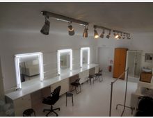 Ein helles Studio mit Schminktischen, die mit integrierten Spiegeln und Leuchtstofflampen ausgestattet sind. Im Raum befinden sich mehrere Stühle, ein Kleiderschrank, eine Dusche, ein Waschbecken und eine Kleiderstange; die Wände und der Boden sind weiß.