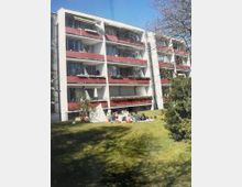 Mehrstöckiges Wohnhaus mit weißer Fassade und durchgehenden Balkonen mit roten Brüstungen. Davor liegt eine große Rasenfläche mit Sträuchern und Bäumen am Rand unter blauem Himmel.