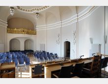 Ein heller Konferenzraum mit gewölbter Decke, goldverzierter Empore und blauen Stühlen, die in Reihen angeordnet sind. Im Vordergrund befinden sich mehrere Rednerpulte mit Mikrofonen, und an den Wänden hängen dekorative Wandlampen.