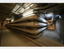 Ein unterirdischer Versorgungstunnel mit mehreren großen Metallrohren, die entlang einer Ziegelwand verlaufen. Die Decke ist mit Kabeltrassen und Beleuchtung ausgestattet, und der Boden besteht aus Beton.