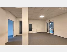 Leerer, rechteckiger Innenraum mit grauem Teppichboden, weißen Wänden und Rasterdecke mit eingebauten Leuchten. Der Raum hat mehrere Türen, darunter eine mit Glaseinsatz und eine verglaste Doppeltür, sowie eine weiße Heizkörperanlage an der Wand.