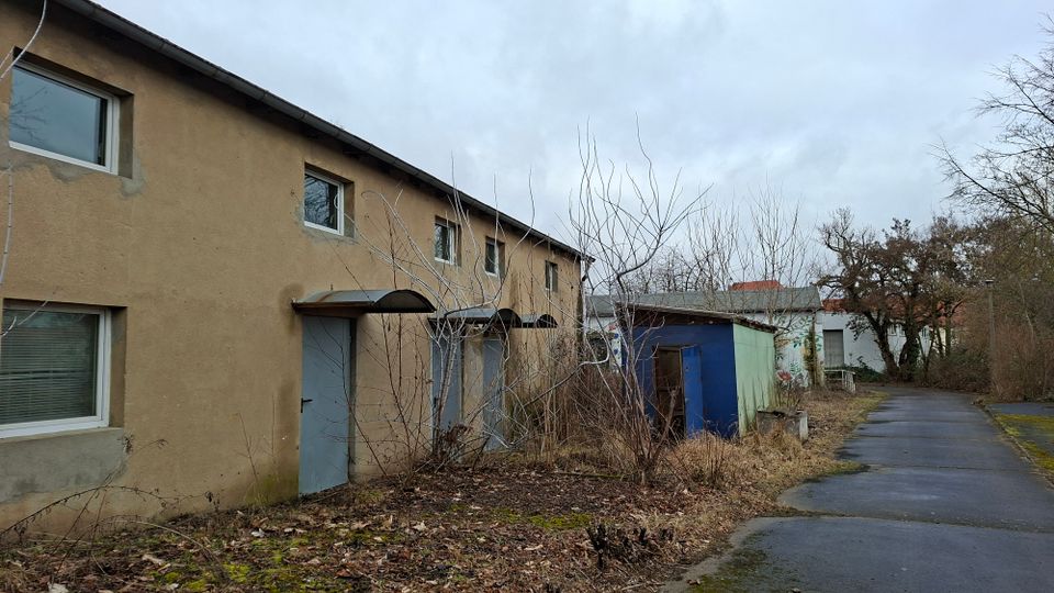 Zweistöckiges, beige verputztes Gebäude mit mehreren Fenstern und grauen Türen unter kleinen Vordächern, daneben ein blaues Nebengebäude und verwilderte Sträucher. Rechts verläuft ein asphaltierter Weg durch das Gelände mit kahlen Bäumen und weiteren Gebäuden im Hintergrund.