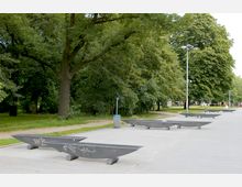 Ein Park mit großen Bäumen auf einer Wiese, neben einem breiten, gepflasterten Weg. Entlang des Weges stehen moderne, graue Sitzbänke in Bootsform sowie Laternen und ein Abfallbehälter.