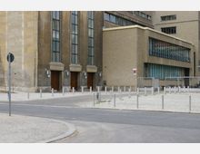 Ansicht eines modernen Gebäudes mit einer Fassade aus Glas und Beton. Links befinden sich große hölzerne Eingangstüren, während rechts ein flacherer Gebäudeteil mit Fenstern und einem Gitterzaun zu sehen ist. Der Vorplatz ist gepflastert und mit Pollern versehen.