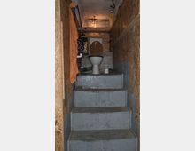 Enge Toilette mit einer Treppe, die zu einer erhöhten Toilettenschüssel führt. Die Wände bestehen aus einer Mischung aus unverputztem Ziegelstein und Sperrholzplatten, mit minimaler Beleuchtung und einfachen Sanitärarmaturen.