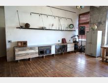 Industriell eingerichteter Raum mit Holzboden, einer rustikalen Holzkommode an der weißen Wand und einer metallischen Hängeregalleiste mit verschiedenen Gegenständen. Im Hintergrund befinden sich ein großer Lautsprecher, ein Metallschrank, eine Weltkugel und eine Fensterfront mit Jalousien.