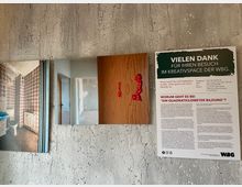 Eine Betonwand mit drei angehängten Objekten: links ein Foto eines alten Badezimmers mit gemusterten Fliesen, in der Mitte ein Bild mit einem roten Türgriff in Form einer Figur auf einer Holztür, und rechts ein Informationsplakat mit Text, Titel „Vielen Dank für Ihren Besuch im Kreativspace der WBG“.