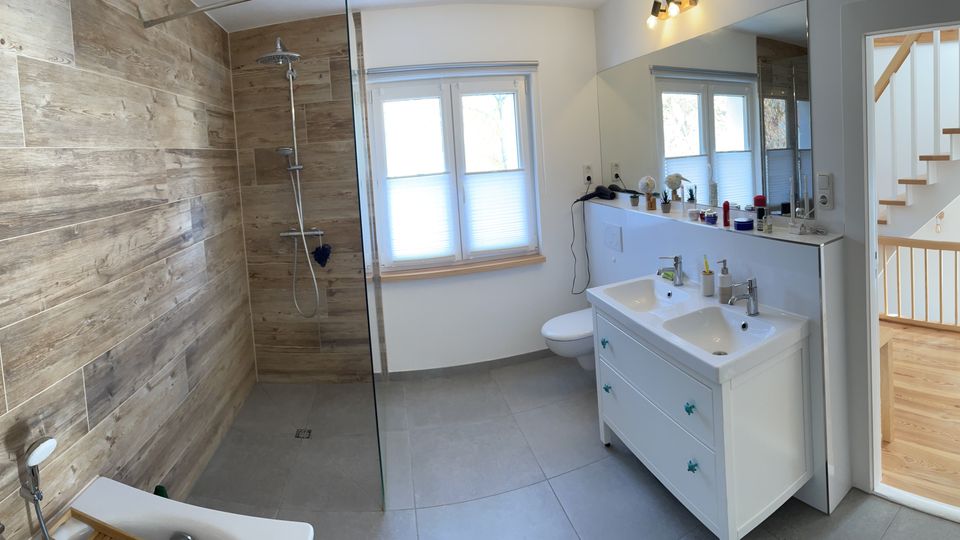 Modernes Badezimmer mit großer begehbarer Dusche hinter Glas, Holzoptik-Wandfliesen und grauen Bodenfliesen. Rechts ein weißer Doppelwaschtisch mit Spiegel, daneben ein Fenster; durch die offene Tür ist ein heller Flur mit Holztreppe sichtbar.