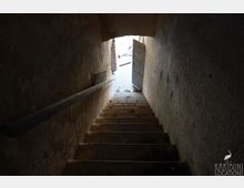 Blick von oben in ein schmales, abgenutztes Treppenhaus mit Betonstufen und rauen Putzwänden, links mit einfachem Handlauf. Unten führt eine halb geöffnete Tür durch einen niedrigen Rundbogen ins helle Außenlicht.