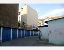 Hinterhof mit blauen Garagentoren, flankiert von einer hohen Steinmauer mit Graffiti und angrenzenden Gebäuden mit Ziegeldächern. Die Wände sind teils verblasst oder beschädigt, und im Hintergrund sind Werbetafeln und Kritzeleien zu sehen.