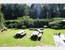 Grüne Wiese mit zwei Holzbänken und Picknicktischen, umgeben von Büschen und Bäumen. Im Hintergrund sieht man eine dichte Hecke, und auf der Wiese liegt ein röhrenförmiges Spielgerät, während rechts ein Steinterrassenbereich beginnt.