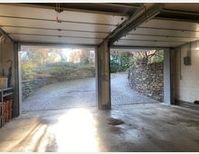 Blick aus einer offenen Doppelgarage mit Betonboden auf eine gepflasterte Auffahrt, die von Natursteinmauern und Sträuchern gesäumt ist. Sonnenlicht fällt durch die Garagentore in den Innenraum.