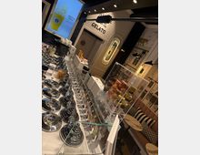 Eisdiele mit moderner Einrichtung: Im Vordergrund eine Glasvitrine mit silbernen Behältern für Eiscreme und dekorativen Eistüten in verschiedenen Farben. Im Hintergrund eine stilisierte Wandgestaltung, gepolsterte Sitzbänke mit Kissen und ein beleuchteter Werbebildschirm.