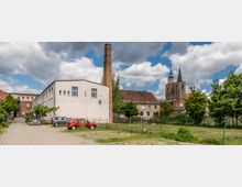 Weite Ansicht eines Stadtviertels mit einer Wiese im Vordergrund, einem modernen weißen Gebäude mit Flachdach und einem hohen Backsteinschornstein in der Mitte. Im Hintergrund sind ältere Gebäude mit Ziegeldächern sowie der Turm einer historischen Kirche mit Spitzdach und Uhr zu sehen.