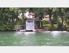 Ein kleines, modernes Steghaus mit glänzender Metallfassade auf einer hölzernen Plattform am Ufer eines Sees, umgeben von dichtem Baumbestand und einem Garten im Hintergrund. Der Steg besitzt eine Leiter, und das Gewässer zeigt eine grünliche Farbe.