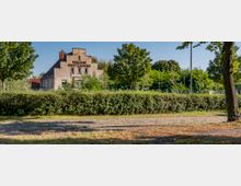 Ehemaliges Industriegebäude mit der Aufschrift "Märkische Kunststeinwerke", umgeben von dichtem Grün und Hecken in einer parkähnlichen Umgebung mit gepflastertem Weg im Vordergrund.