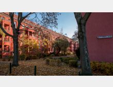 Wohnanlage mit roten Backsteinfassaden und Balkonreihen, umgeben von einem begrünten Hof mit Bäumen, Sträuchern und herbstlichem Laub. Ein Straßenschild mit der Aufschrift „Am Steinberg 104-104E“ ist an der Wand eines Gebäudes angebracht.