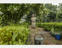 Ein gepflegter Garten mit einer niedrigen Hecke, blühenden Rosensträuchern und einer Marmorbüste auf einem Sockel. Im Vordergrund steht eine Gießkanne, und im Hintergrund sind Bäume und ein grünes Metallgittertor zu sehen.