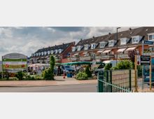 Straßenszene mit einem Wochenmarkt in einer Wohngegend; im Hintergrund stehen Reihenhäuser mit dunklen Dächern und Balkonen, während im Vordergrund Verkaufsstände unter grünen Markisen sowie ein Schild mit der Aufschrift "Werderaner Frischemarkt" zu sehen sind.