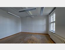 Leerer Raum mit Holzdielenboden, weißen Wänden und einer großen Fensterfront auf der rechten Seite, die natürliches Licht hereinlässt. An der Decke hängen lange Leuchtstofflampen, und unter dem Fenster befindet sich ein Heizkörper.