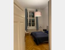 Ein schlicht eingerichtetes Schlafzimmer mit Holzboden, einem Bett mit dunkelblauer Bettdecke, einem Sessel neben einem Heizkörper unter einem großen Fenster und einer hängenden Deckenleuchte mit Lampenschirm. Die Fenster sind mit weißen Gardinen und blauen Vorhängen dekoriert.