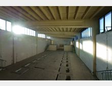 Innenraum einer leerstehenden Industriehalle mit hoher Decke und sichtbarer Trägerkonstruktion. Große Fensterreihen an beiden Seitenwänden lassen Tageslicht hinein, während der Boden von rechteckigen Öffnungen durchzogen ist, die mögliche Wartungsgruben oder Schächte darstellen.